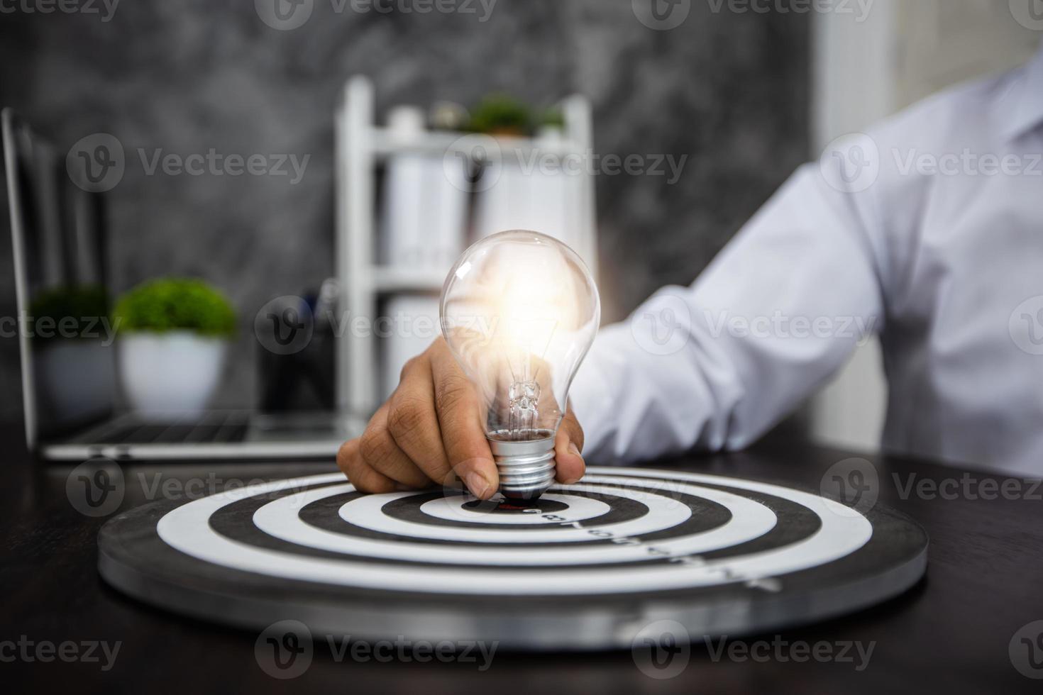 Geschäftsmann, der beleuchtete Glühbirne auf einer Dartscheibe auf einem schwarzen Schreibtisch hält foto