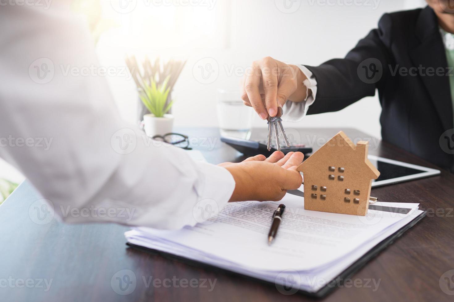 Geschäftsmann, der einer Person neben einem Tablet, einem Musterhaus und einem Vertrag Schlüssel übergibt foto