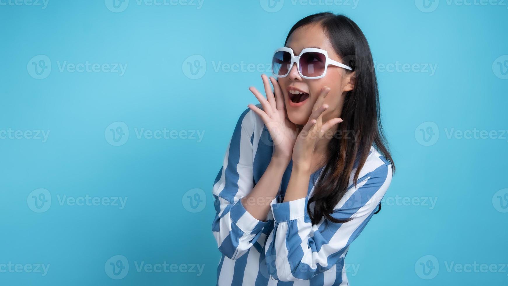 asiatische Frau mit weißer Sonnenbrille mit Händen neben dem Mund, der in Richtung Kopierraum auf blauem Hintergrund gestikuliert foto