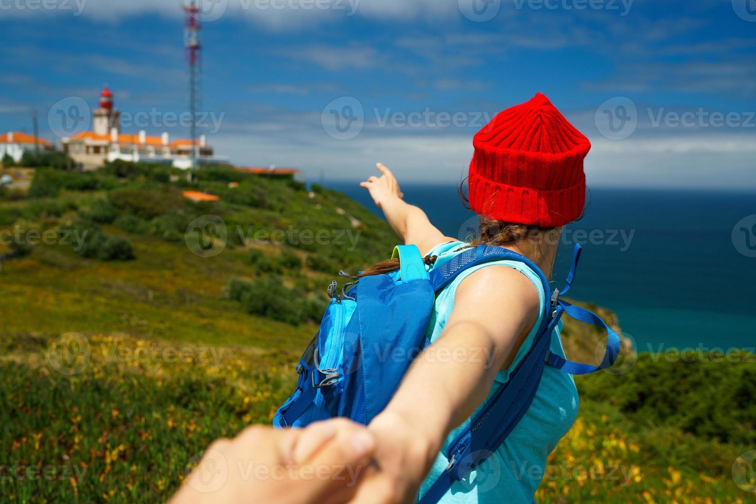 Folgen mich - - glücklich jung Frau im ein rot Hut und mit ein Rucksack hinter ihr zurück ziehen Leute Hand. Hand im Hand Gehen zu das Leuchtturm foto