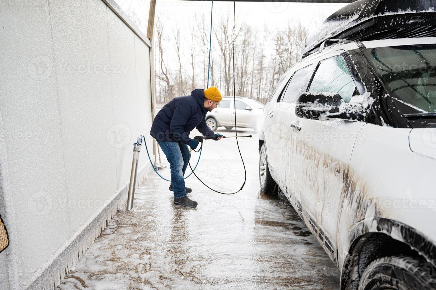 Mann wäscht Hochdruckwasser amerikanisches SUV-Auto mit Dachgepäckträger bei Selbstbedienungswäsche bei kaltem Wetter. foto