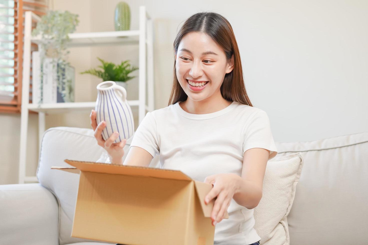 glücklich aufgeregt, asiatisch jung Frau, Mädchen Kunde Sitzung auf Sofa beim heim, Öffnung und Auspacken Karton Box Karton Paket nach Kauf Bestellung gegenwärtig, Einkaufen online, Lieferung Bedienung Konzept. foto