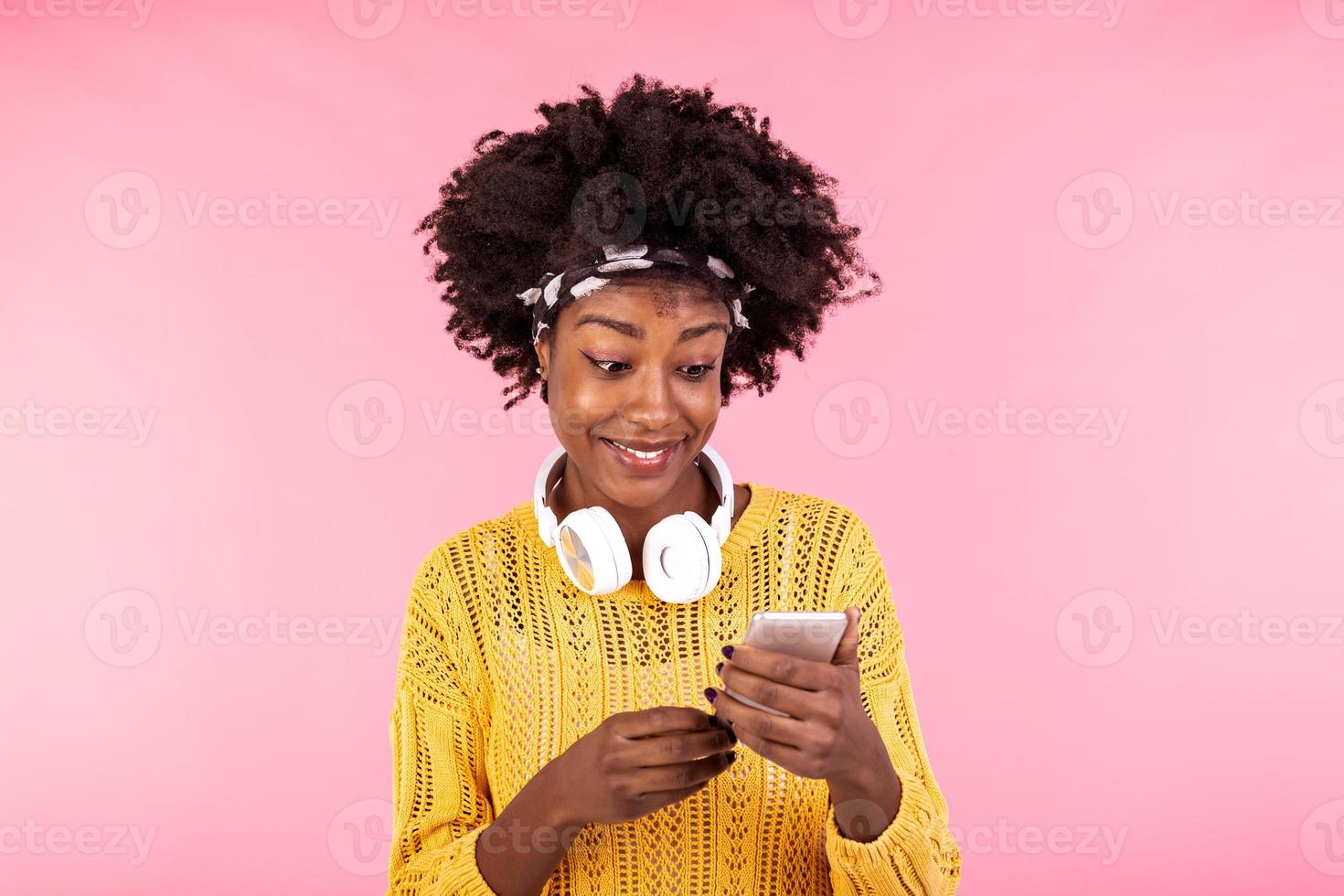 afrikanisch amerikanisch Frau tragen Kopfhörer und lächelnd. jung Frau mit natürlich lockig Haar halten und suchen beim Handy, Mobiltelefon Telefon. foto