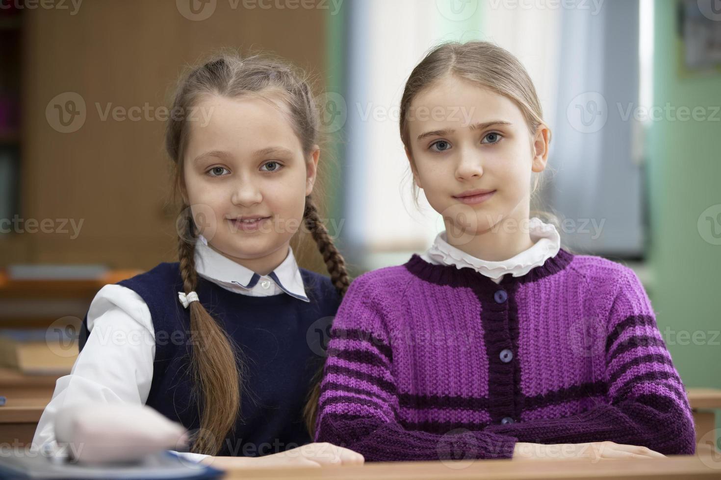 zwei Schulmädchen sitzen beim ein Schreibtisch im das Klassenzimmer und aussehen beim das Kamera. foto