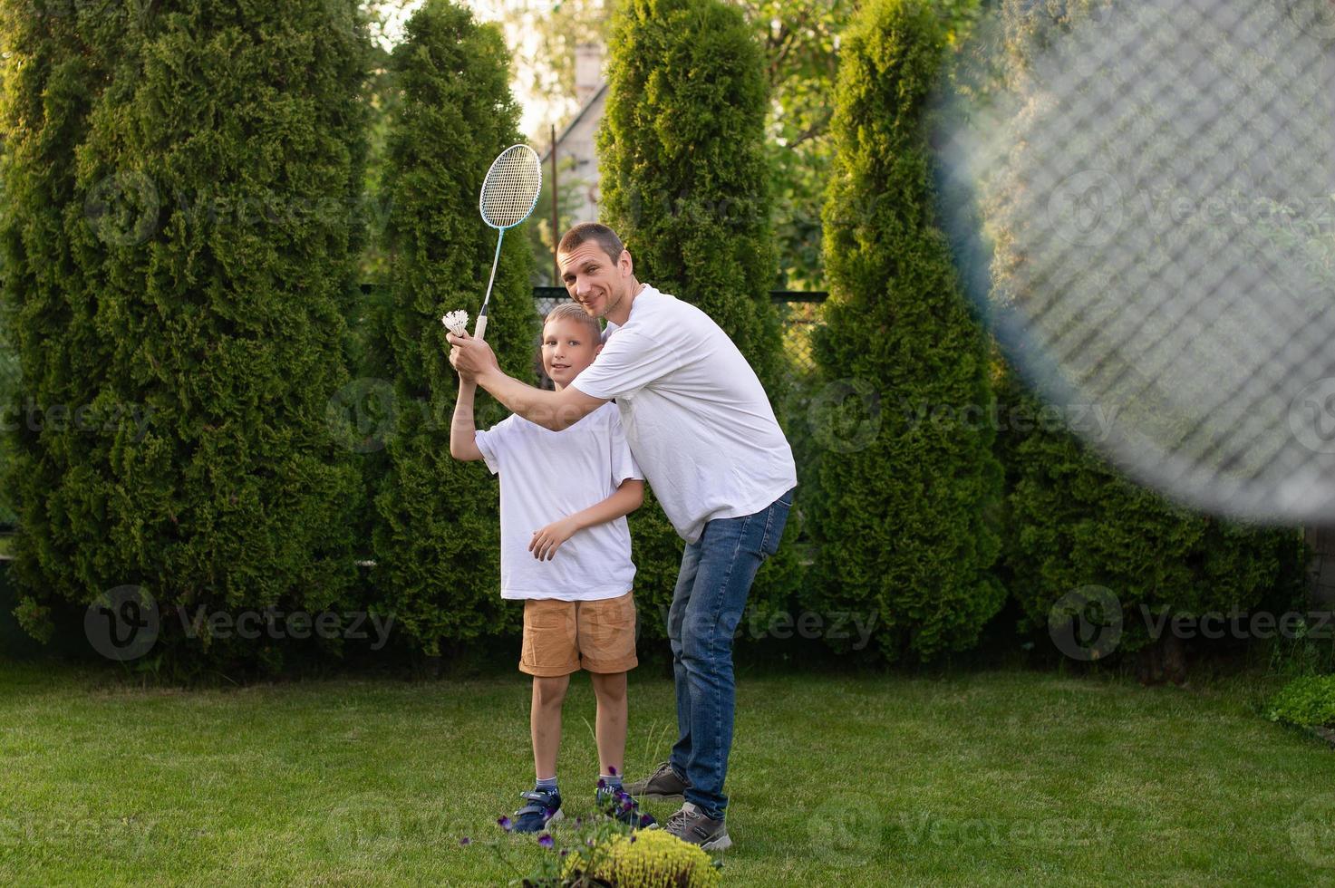 ein heiter Papa auf das Straße unterrichtet seine Sohn zu abspielen Badminton im ein Weiß T-Shirt foto