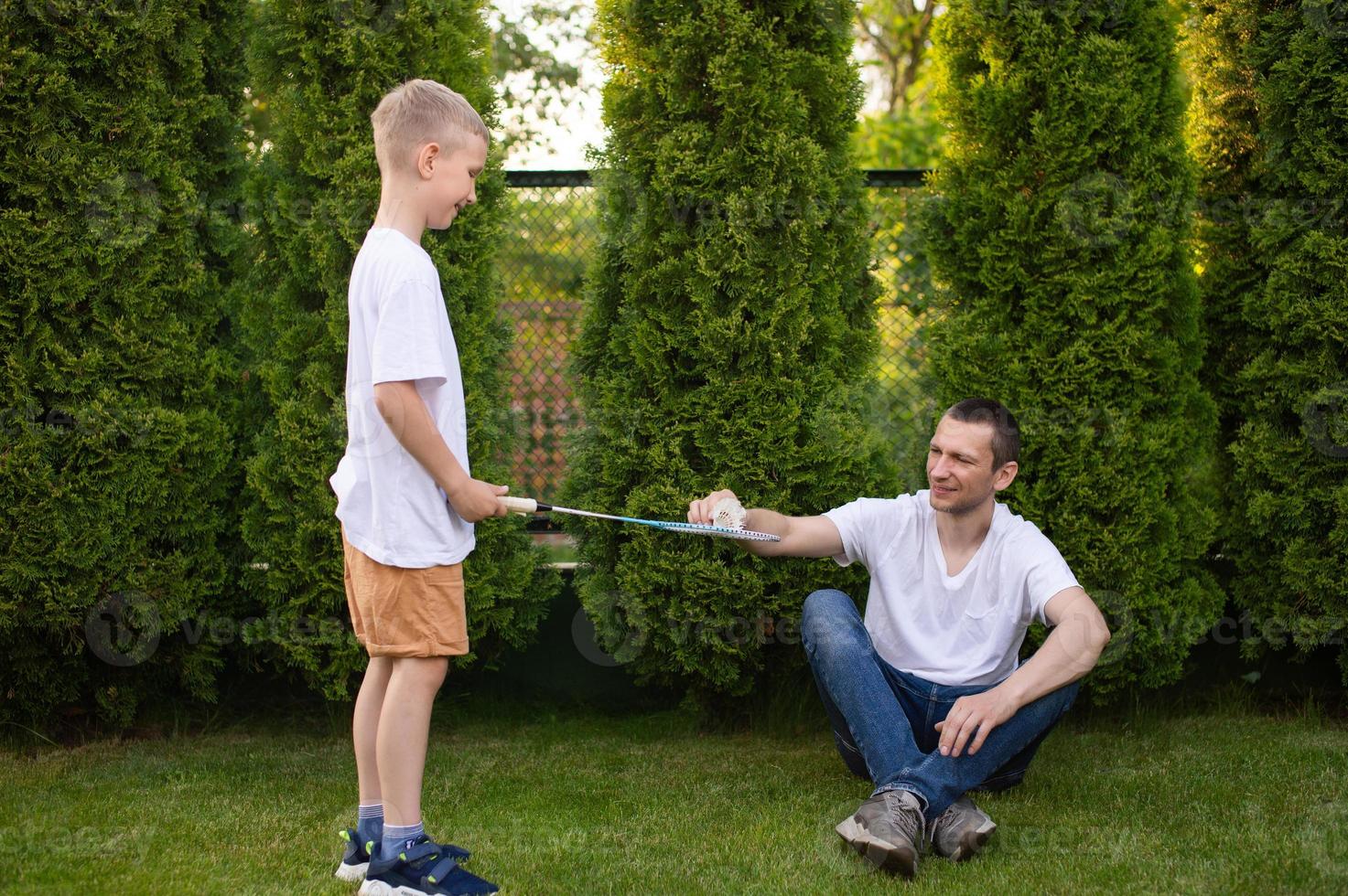 ein heiter Papa auf das Straße unterstützt seine Sohn. ein Junge ist spielen Badminton im ein Weiß T-Shirt foto