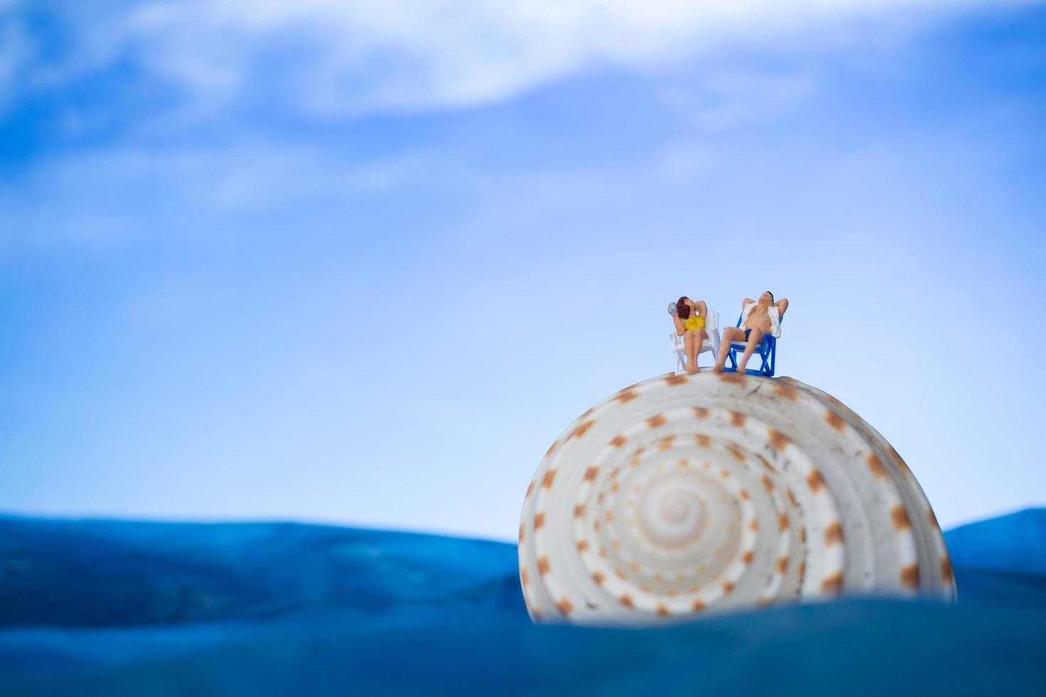 Miniaturmenschen, die auf einer Muschel mit einem Hintergrund des blauen Himmels, Sommerferienkonzept sonnenbaden foto