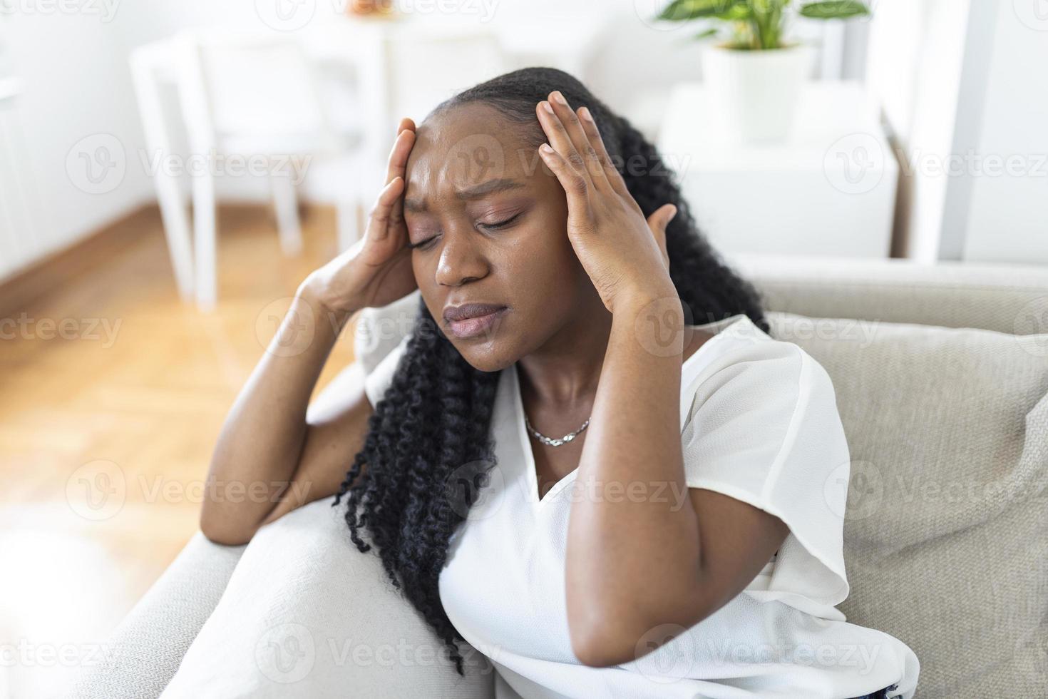 Porträt von ein jung schwarz Mädchen Sitzung auf das Couch beim Zuhause mit ein Kopfschmerzen und Schmerz. schön Frau Leiden von chronisch Täglich Kopfschmerzen. traurig Frau halten ihr Kopf weil Sinus Schmerzen foto