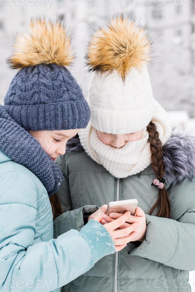 zwei Mädchen im warm Mäntel und Hüte schreiben ein Text Botschaft auf ein Smartphone im ein schneebedeckt Winter Park. Lebensstil verwenden von Technologie. Vertikale Aussicht foto