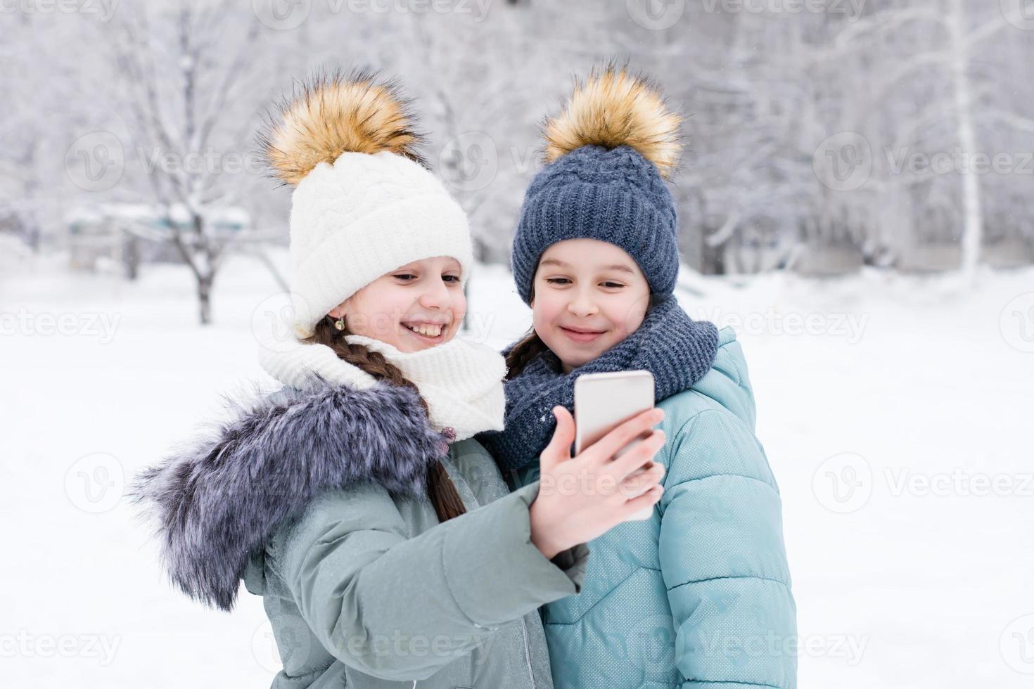 zwei lächelnd Mädchen im warm Kleider machen ein Video Anruf auf ein Smartphone im ein schneebedeckt Winter Park. Lebensstil verwenden von Technologie foto