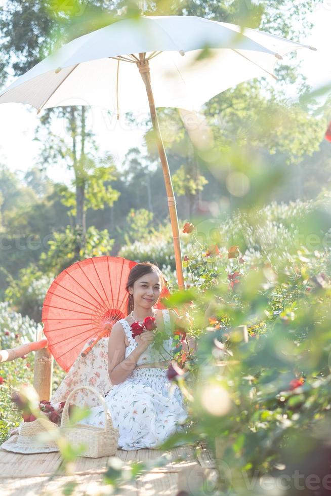 jung asiatisch Frau tragen ein Weiß Kleid posiert mit ein Rose im Rose Garten foto