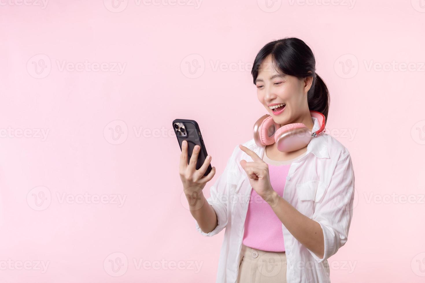 Porträt jung attraktiv asiatisch Frau glücklich Lächeln mit Smartphone mit Kopfhörer, Kopfhörer isoliert auf Rosa Studio Hintergrund. ziemlich weiblich Person mit Handy, Mobiltelefon Telefon. Musik- online Lebensstil Konzept. foto