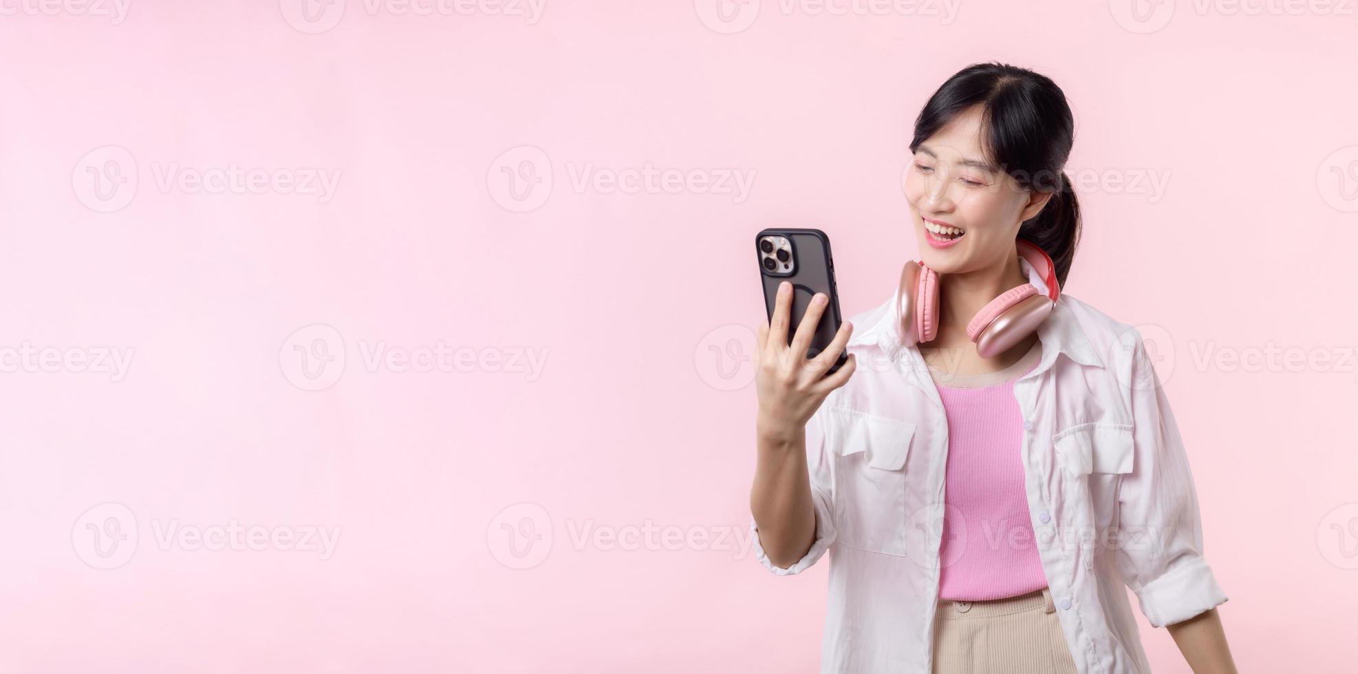 Porträt jung attraktiv asiatisch Frau glücklich Lächeln mit Smartphone mit Kopfhörer, Kopfhörer isoliert auf Rosa Studio Hintergrund. ziemlich weiblich Person mit Handy, Mobiltelefon Telefon. Musik- online Lebensstil Konzept. foto