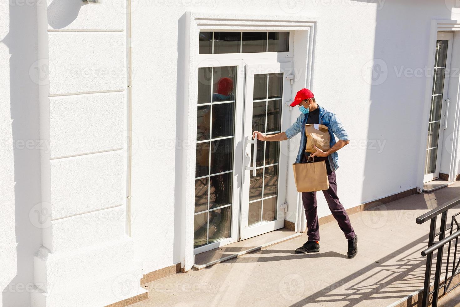 Lieferung Mann halten Papier Tasche mit Essen auf Weiß Eingang von Haus Hintergrund , Essen Lieferung Mann im schützend Maske foto