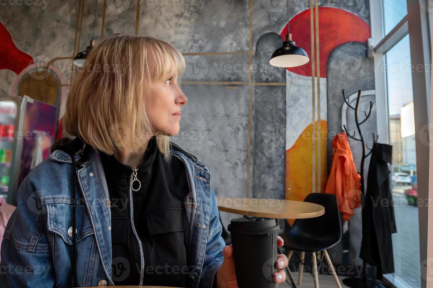 ein blond Frau im ihr vierziger Jahre sitzt allein im ein Cafe, ruht, Getränke Kaffee und sieht aus aus das Fenster beim das Stadt Straße foto