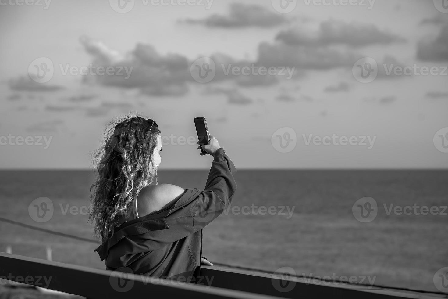 ein schön blond Frau mit lange wellig Haar steht auf ein Brücke und schießt ein Sonnenuntergang beim das Meer auf ihr Telefon, schwarz und Weiß Foto