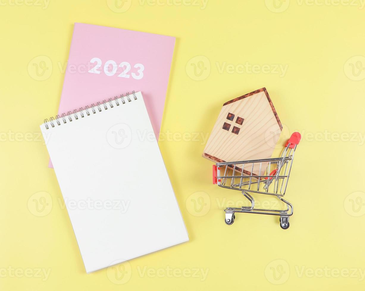 eben Layout von hölzern Haus Modell- im Einkaufen Wagen oder Einkaufen Wagen mit leer Seite Notizbuch und Rosa Tagebuch oder 2023 auf Gelb Hintergrund mit Kopieren Raum, Zuhause Kauf planen Konzept. foto