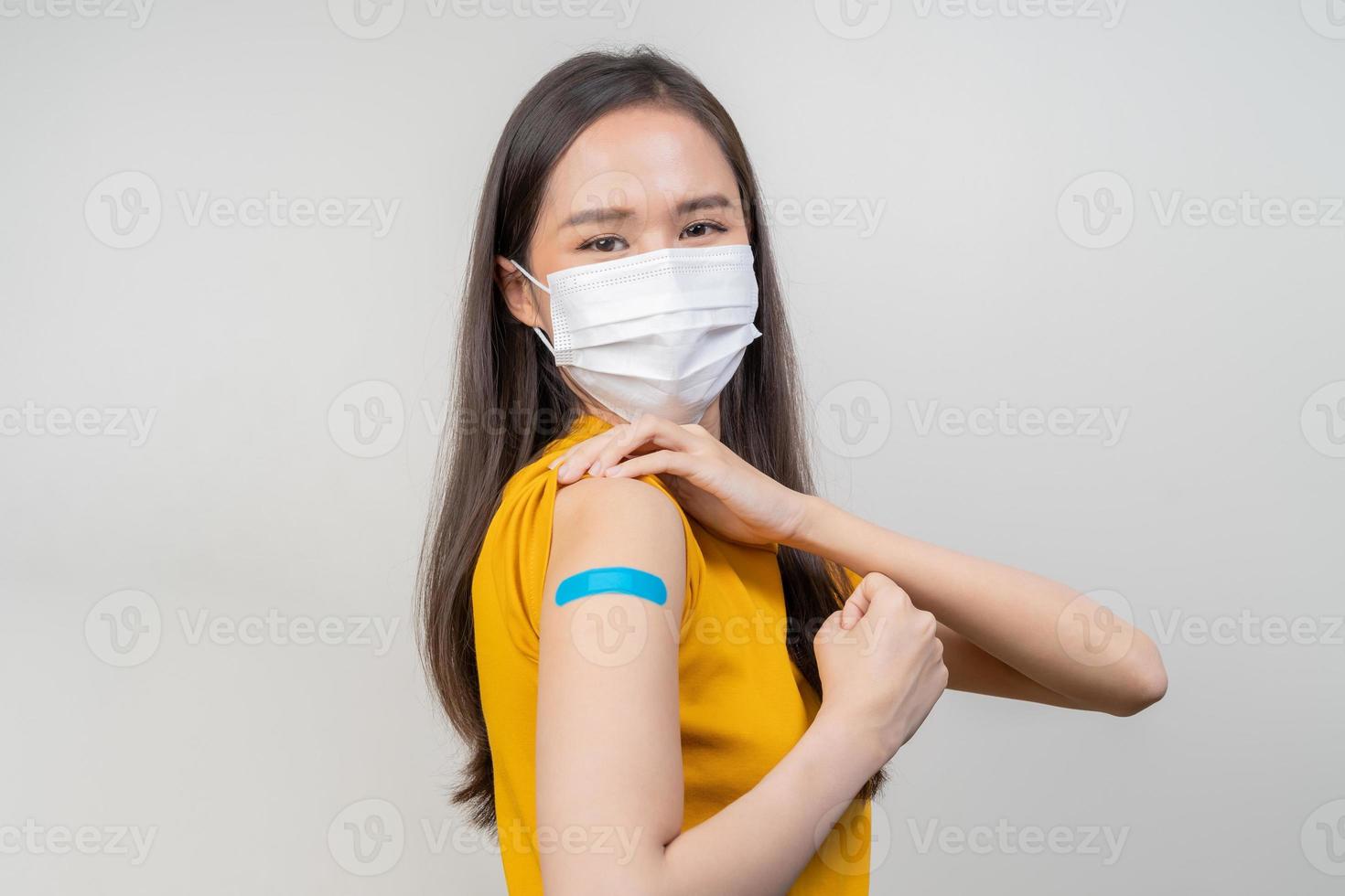 Porträt von Gesundheit Pflege asiatisch jung Frau tragen Gesicht Maske schützen, bekommen Impfstoff von COVID-19, Grippe im Gelb T-Shirt, zeigen Arm auf Blau Bandage, isoliert auf Natur Hintergrund, Kopieren Raum. foto