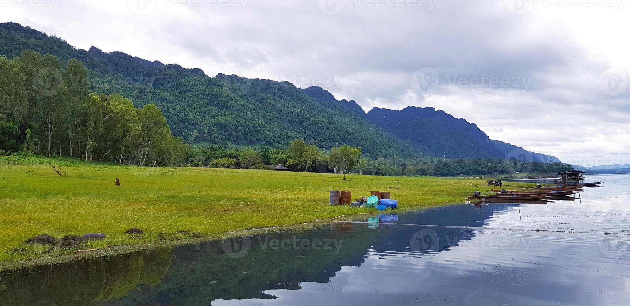schön von Landschaft Sicht. lange Schwanz Boot geparkt oder schwebend auf das Wasser mit Grün Gras, Baum, groß Berg und Wolke Himmel Hintergrund beim Srinakarin See, Kanchanaburi, Thailand. Natur Hintergrund foto