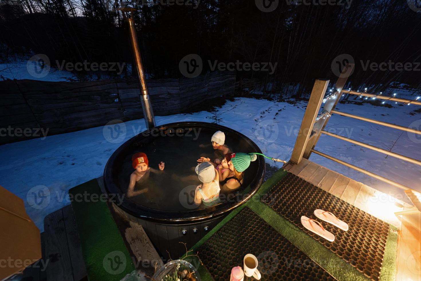 Familie genießen Baden im hölzern Fass heiß Wanne im das Terrasse von das Hütte. skandinavisch Badewanne mit ein Kamin zu brennen Holz und Hitze Wasser. foto