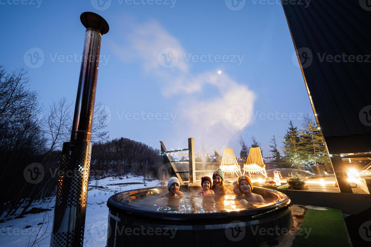 Familie genießen Baden im hölzern Fass heiß Wanne im das Terrasse von das Hütte. skandinavisch Badewanne mit ein Kamin zu brennen Holz und Hitze Wasser. foto