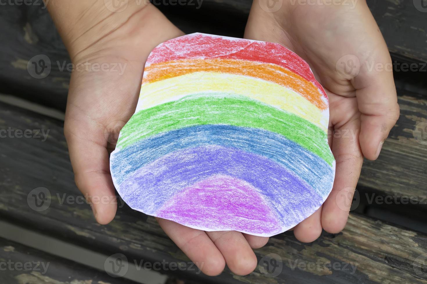 das Hände von ein klein Kind sind sanft halten ein Regenbogen Stück von Papier gezeichnet mit farbig Bleistifte. foto