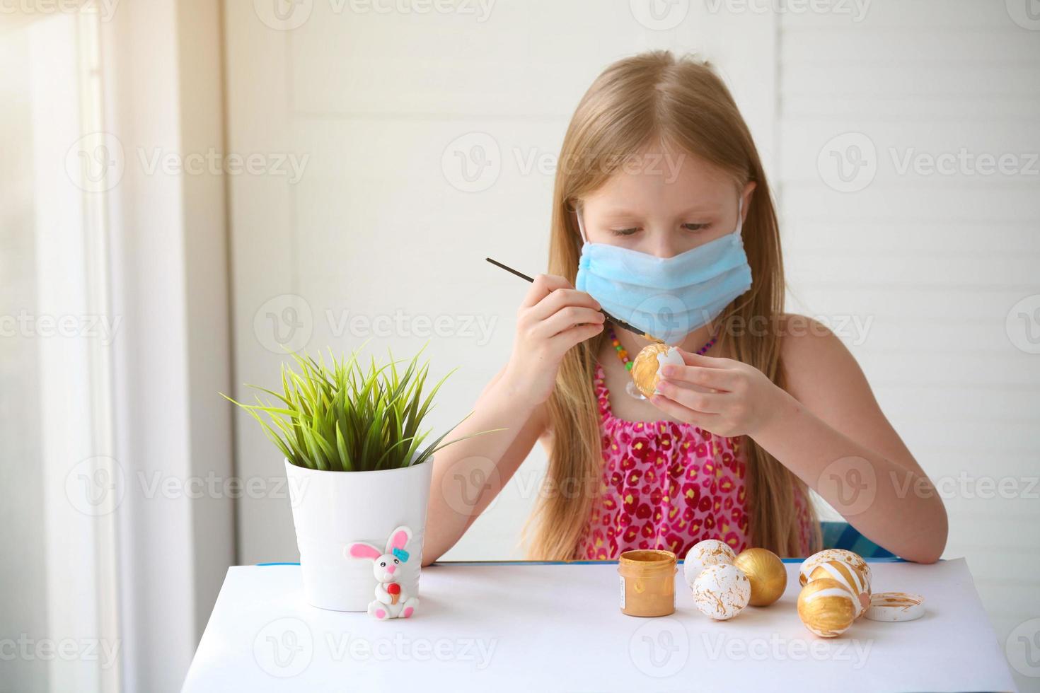 wenig Mädchen im ein medizinisch Maske Farben Ostern Eier mit ein Muster. foto