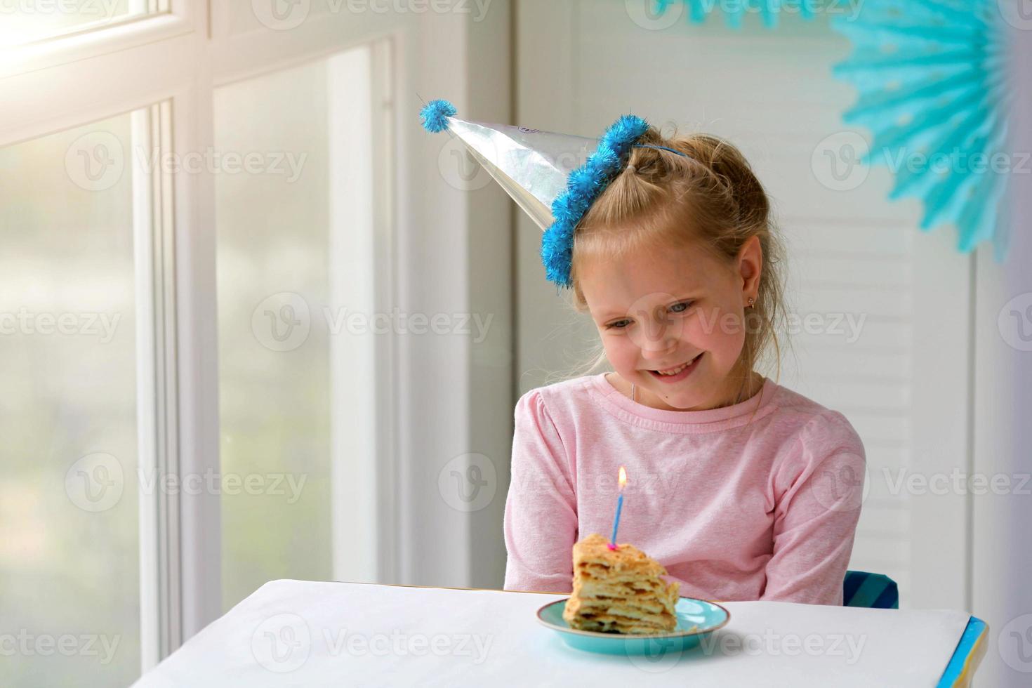 glücklich wenig Mädchen sitzt im ein Hut im Vorderseite von ein Stück von Kuchen und ein Kerze. Geburtstag beim heim. foto