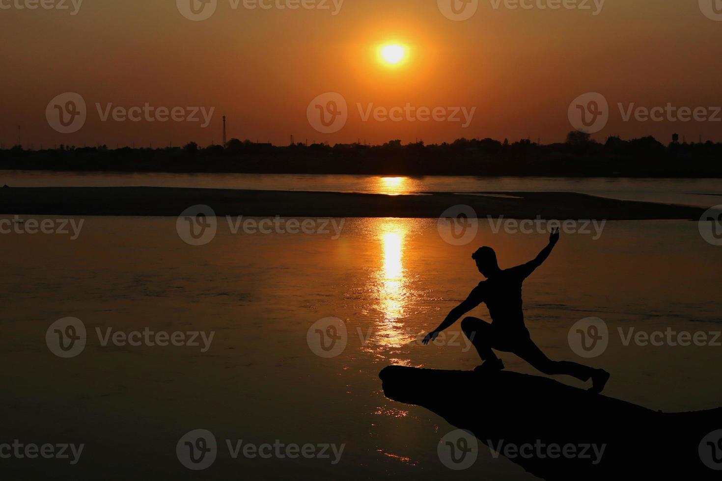 Silhouette von ein Mann tun Yoga auf das Bank von das Fluss beim Sonnenuntergang. gesund Konzept. foto