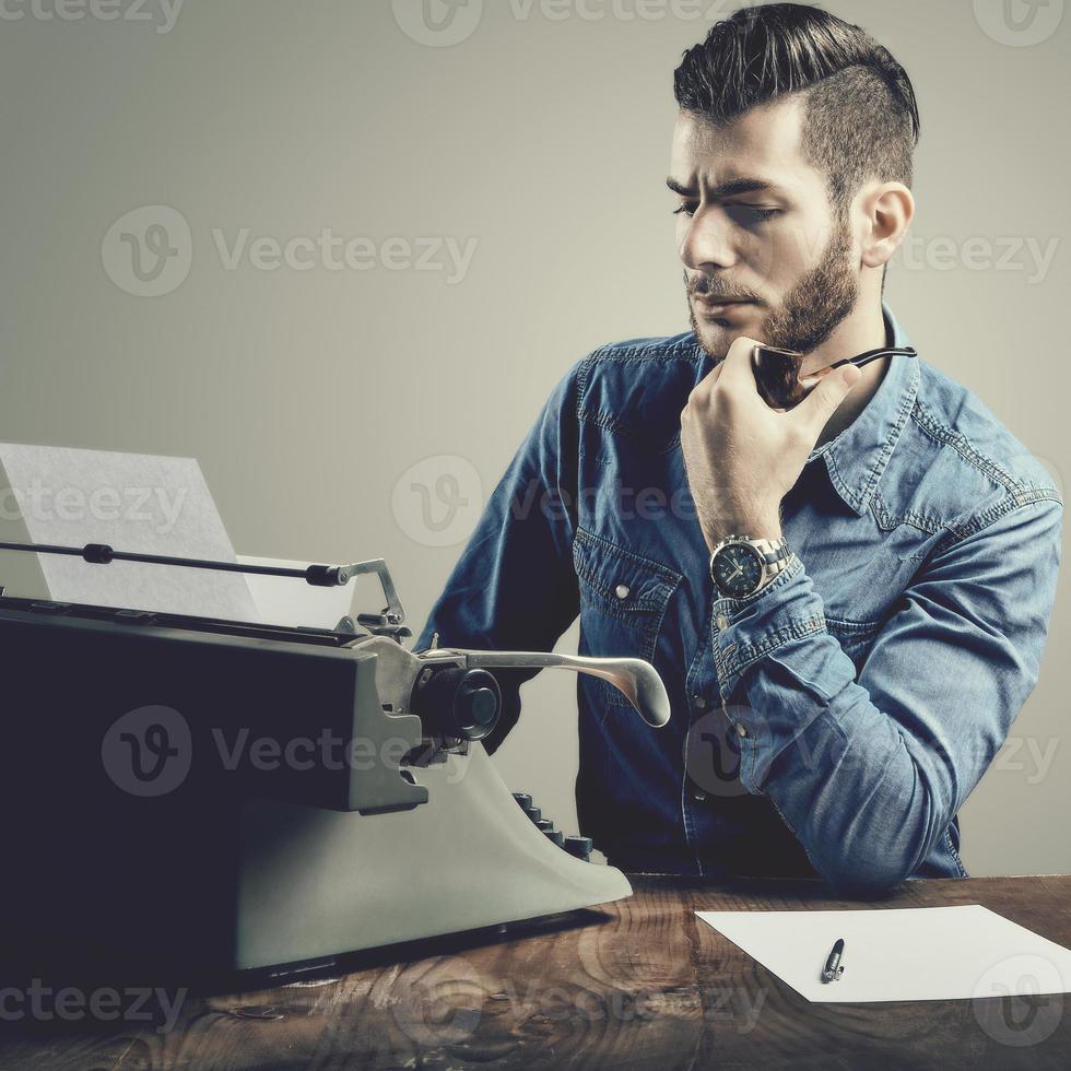 junger Mann mit Bart und Schnurrbart an der Schreibmaschine, die seine Pfeife raucht foto