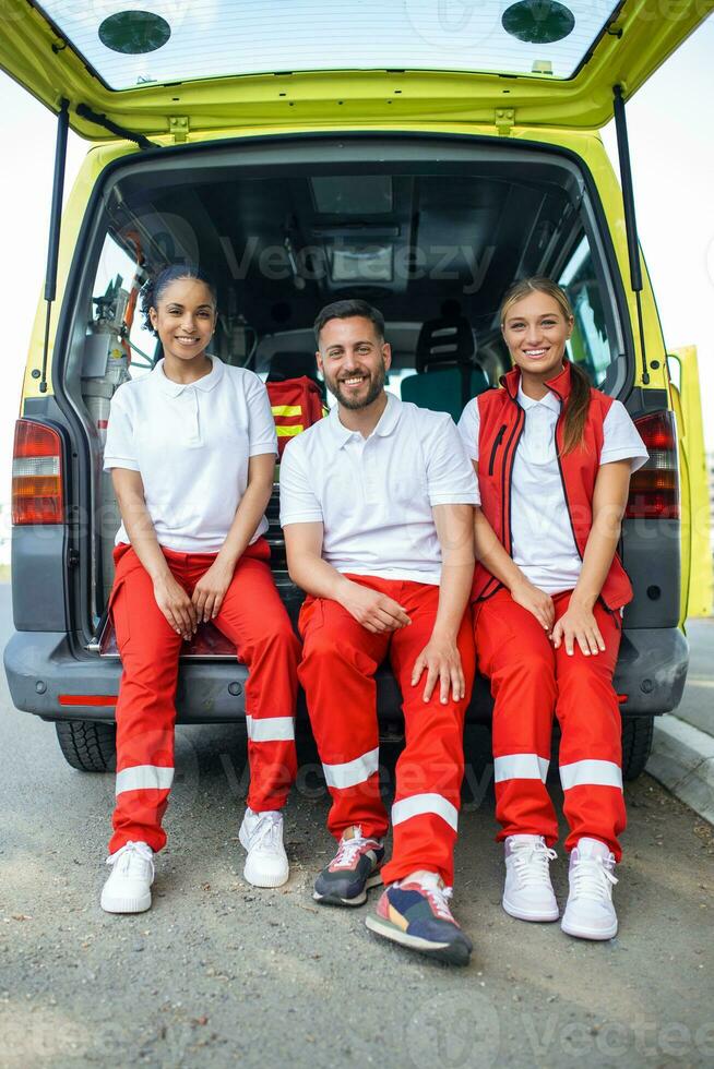 multiethnisch Gruppe von Sanitäter Stehen beim das Seite von ein Krankenwagen mit öffnen Türen. ihr Mitarbeiter Tragen ein medizinisch Trauma Tasche. Sie sind lächelnd beim das Kamera. foto