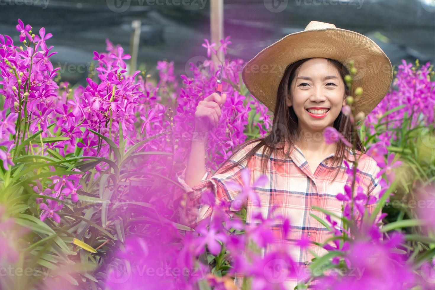 jung weiblich Gärtner tragen ein Plaid Hemd und tragen ein Hut. Hände halten Schere zum Schneiden Orchideen und lächeln. Neu Generation Bauer, Gärtner Konzept. foto