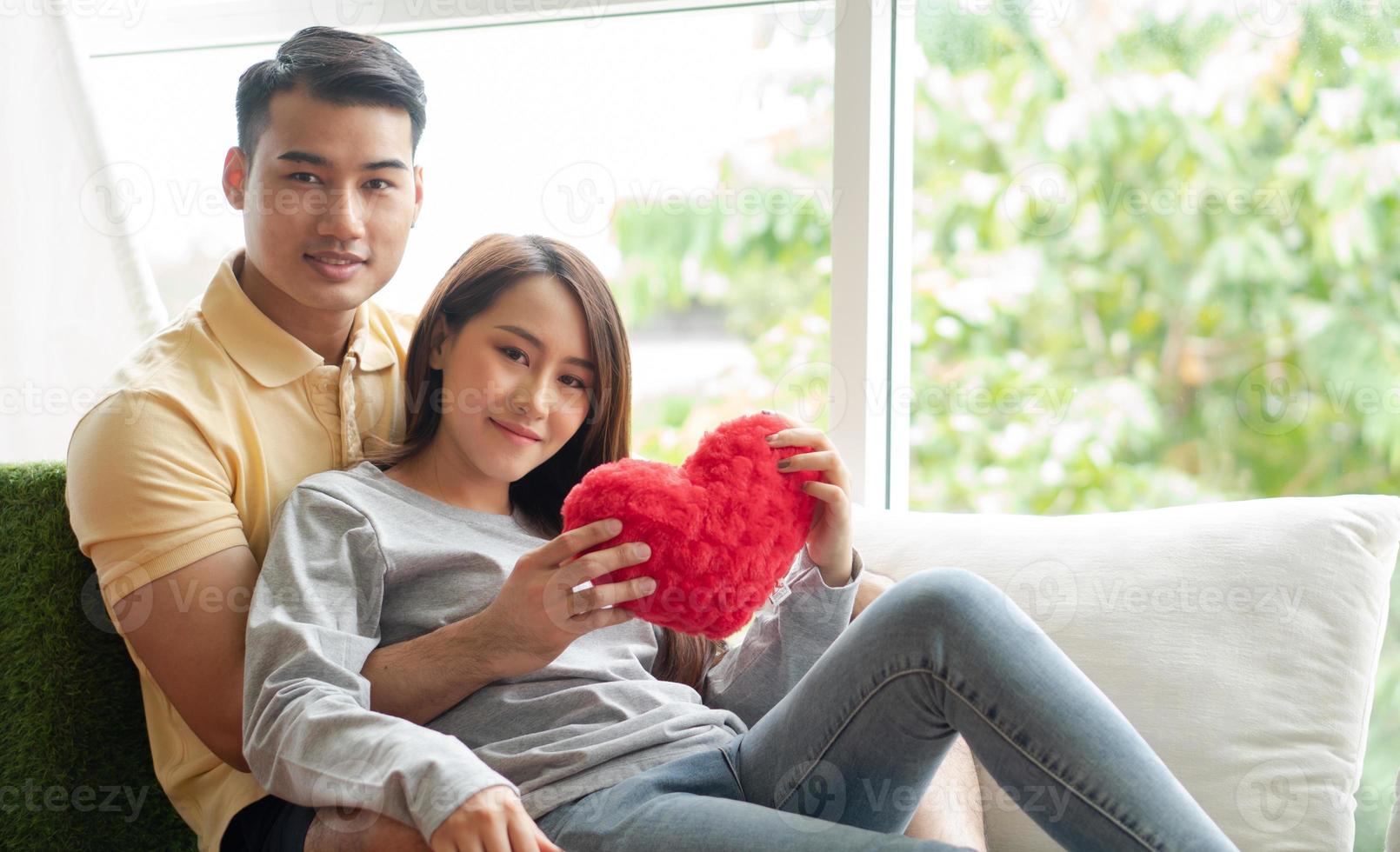 glücklich Paar Sitzung auf das Sofa und Sein ein Mann Umarmen seine Freundin mit Liebe im das Leben Zimmer und Lächeln und halten ein rot Herz. Konzept von romantisch auf Valentinstag Tag. Vorschlag und Ehe foto