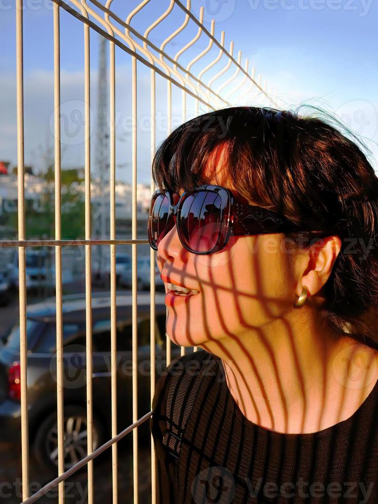 abspielen von Abend Beleuchtung Schatten auf das Gesicht, Porträt von ein weiblich asiatisch im schwarz Kleid tragen Sonnenbrille, Vertikale Weiß Metall Zaun Schatten Gießen auf ihr Gesicht, Sonnenuntergang foto
