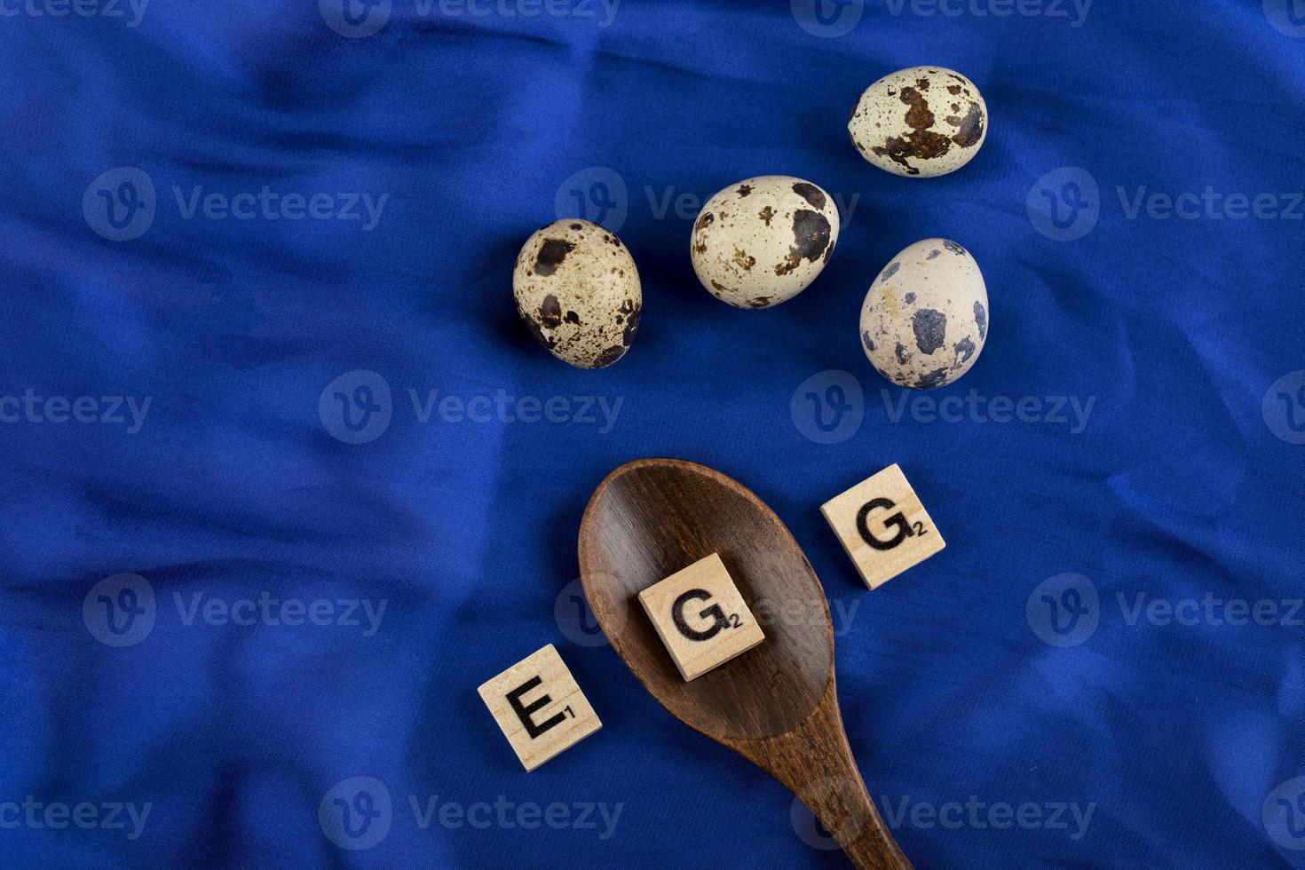 Wachteleier mit einem Holzlöffel und Holzbuchstaben, die das Wort Ei auf einem blauen Satintuch buchstabieren foto