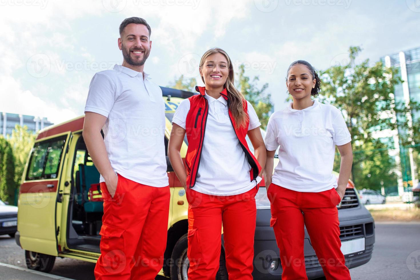 drei gemischtrassig Sanitäter Stehen im Vorderseite von Krankenwagen Fahrzeug, Tragen tragbar Ausrüstung foto