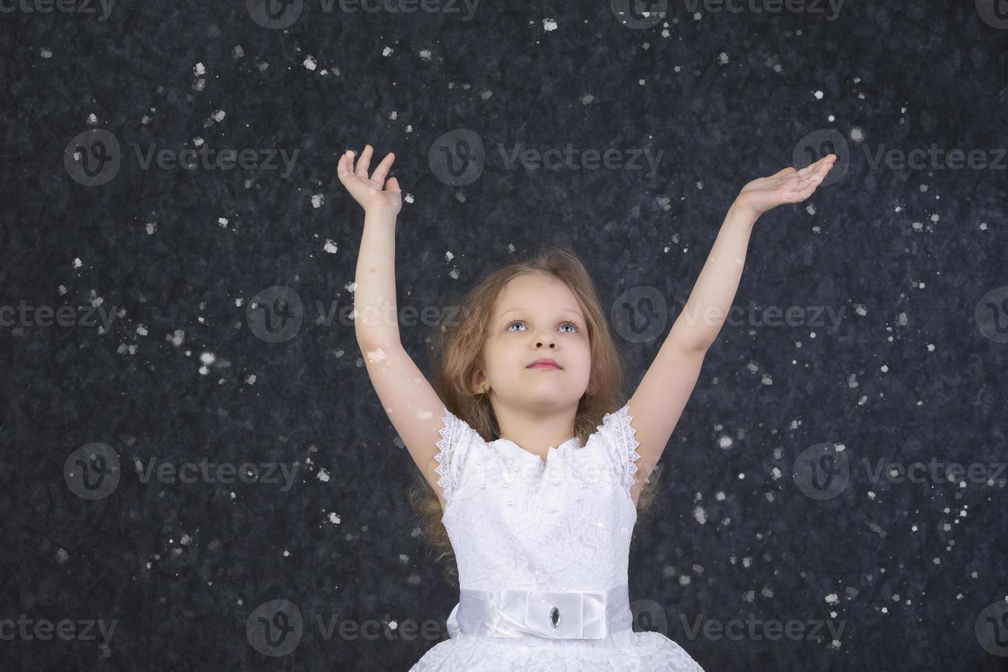 schön wenig Mädchen im ein Weiß Kleid mit angehoben Waffen unter fliegend Schneeflocken. foto