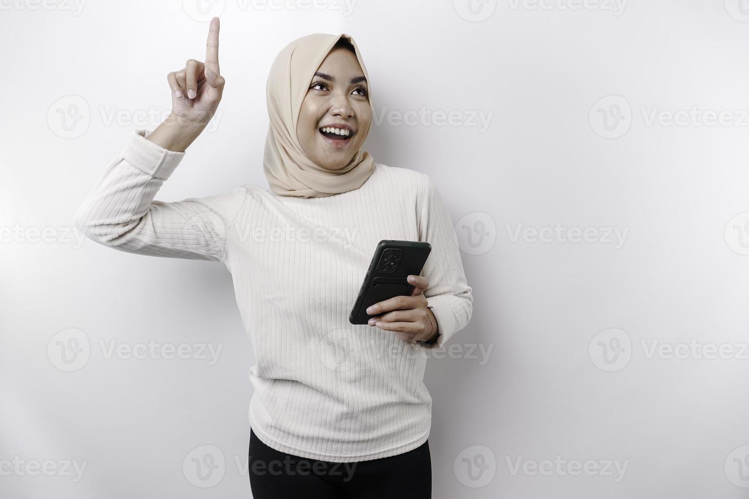 Aufgeregte asiatische Frau, die Hijab trägt und auf den Kopierbereich auf ihr zeigt, während sie ihr Telefon hält, isoliert durch weißen Hintergrund foto