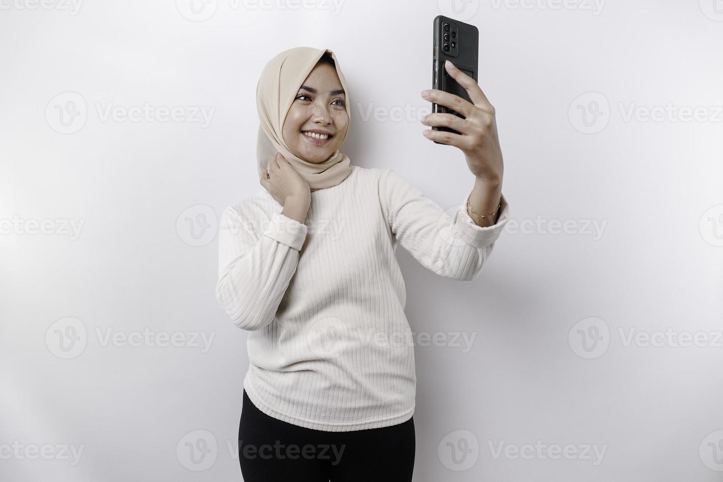 ein glücklich asiatisch Muslim Frau tragen ein Kopftuch, halten ihr Telefon, isoliert durch Weiß Hintergrund foto