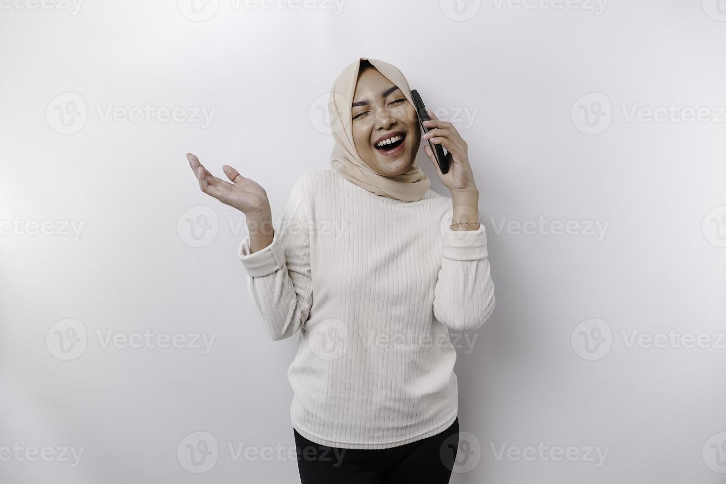 Aufgeregte asiatische Frau mit Hijab, die auf den Kopierbereich neben ihr zeigt, während sie ihr Telefon hält, isoliert durch weißen Hintergrund foto