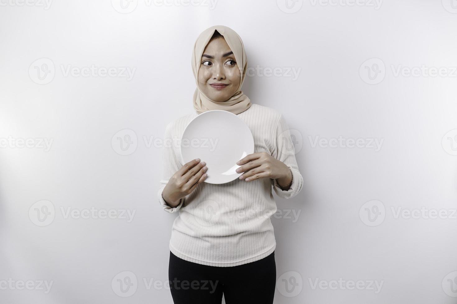 ein asiatisch Muslim Frau ist Fasten und hungrig und halten ein Teller während suchen beiseite Denken Über Was zu Essen foto