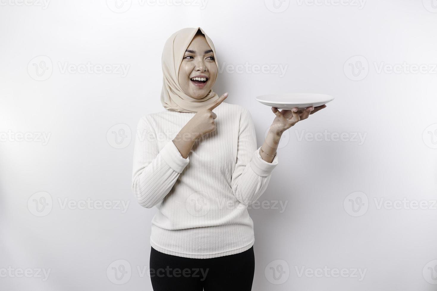 ein lächelnd asiatisch Muslim Frau ist Fasten und hungrig und halten und zeigen zu ein Teller foto