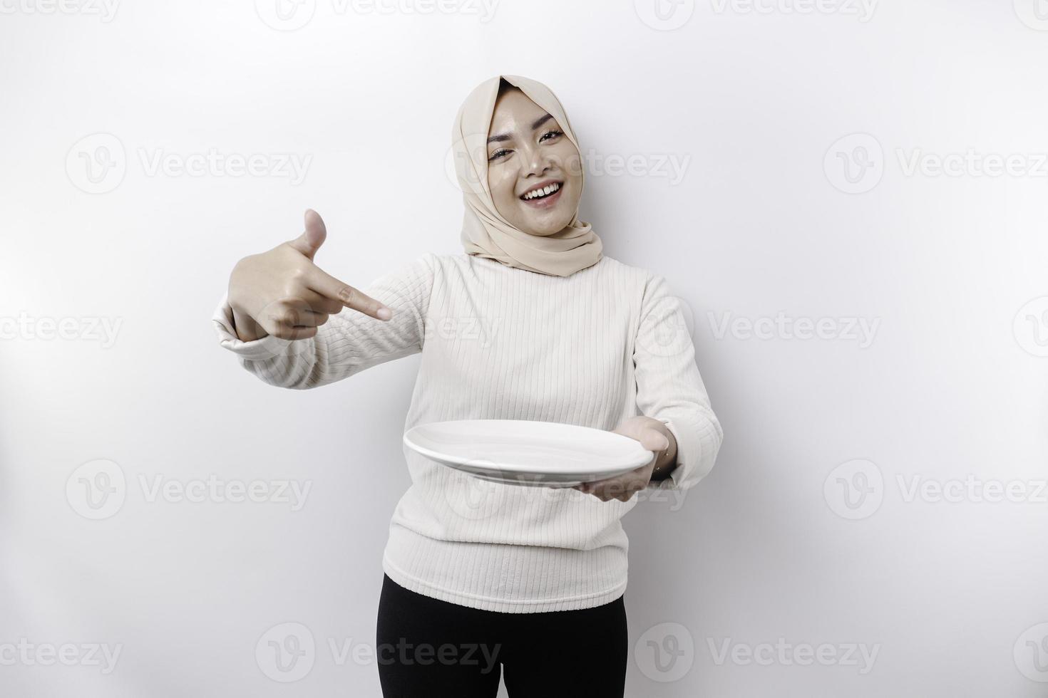 ein lächelnd asiatisch Muslim Frau ist Fasten und hungrig und halten und zeigen zu ein Teller foto