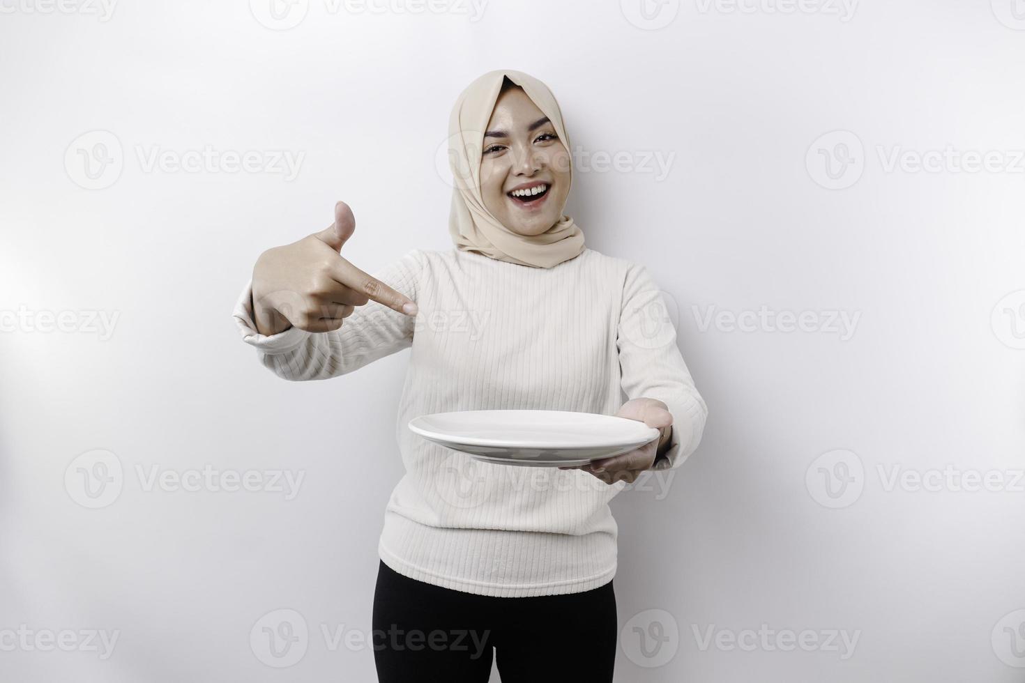 ein lächelnd asiatisch Muslim Frau ist Fasten und hungrig und halten und zeigen zu ein Teller foto