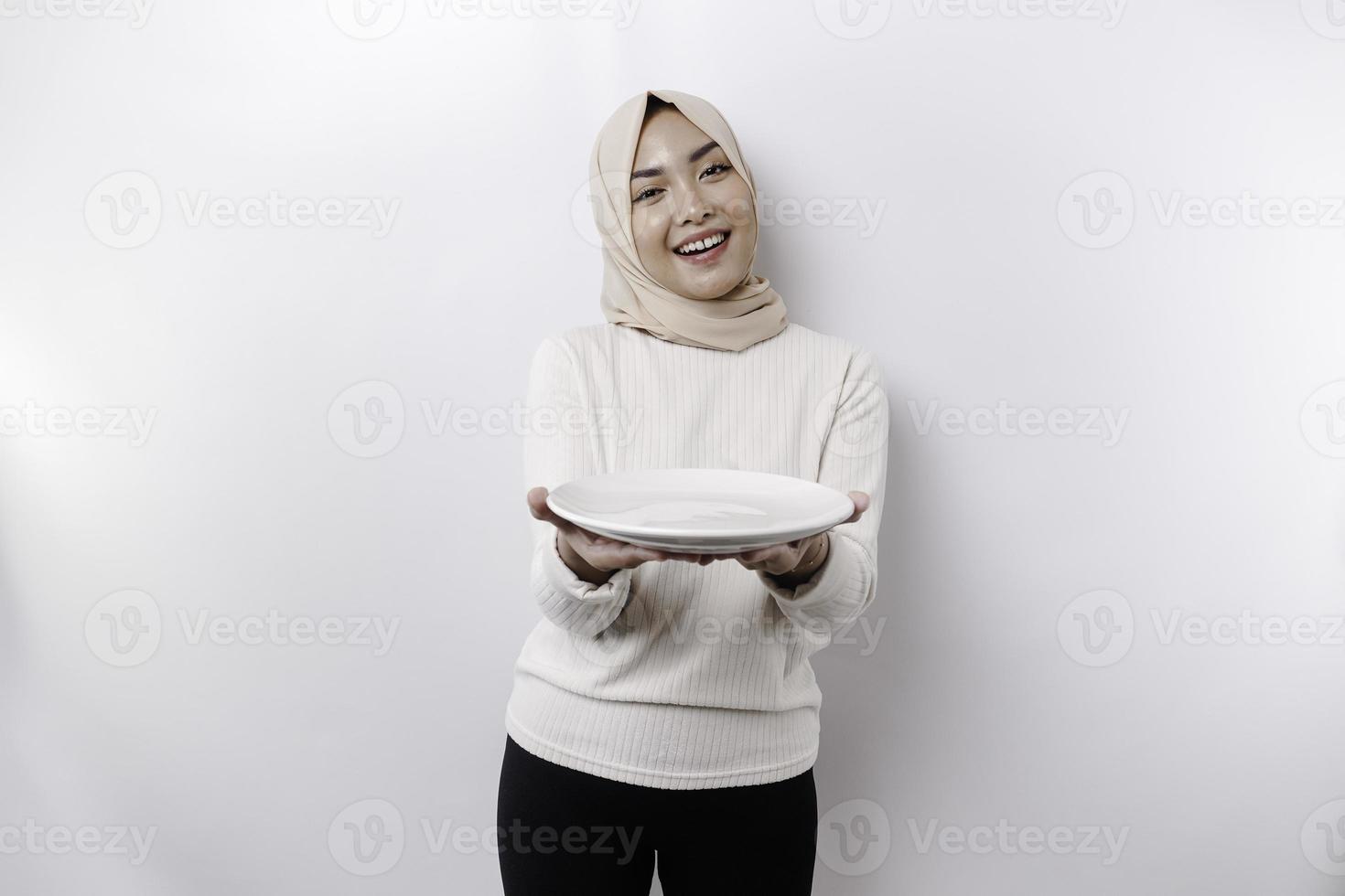 ein lächelnd asiatisch Muslim Frau ist Fasten und hungrig und halten und zeigen zu ein Teller foto