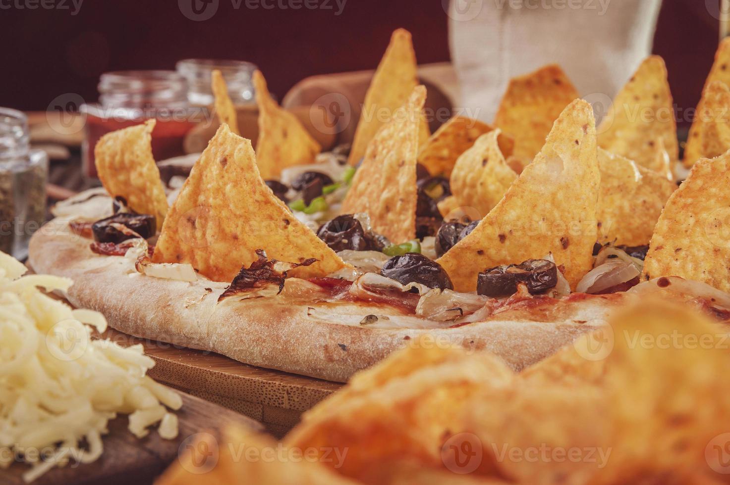 Pizza mit Mozzarella, Zwiebeln, Peperoni, schwarzen Oliven, grünem Paprika, Nachos und Oregano foto