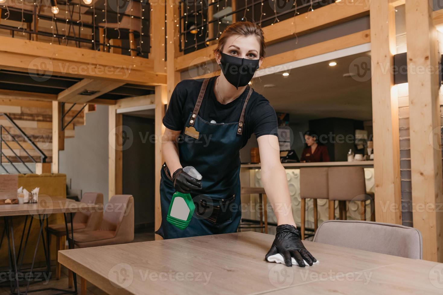 Eine freundliche Kellnerin, die eine schwarze medizinische Gesichtsmaske und medizinische Einweghandschuhe trägt, hält eine Flasche mit Desinfektionsmittel und Putztischen mit einem Lappen in einem Restaurant foto