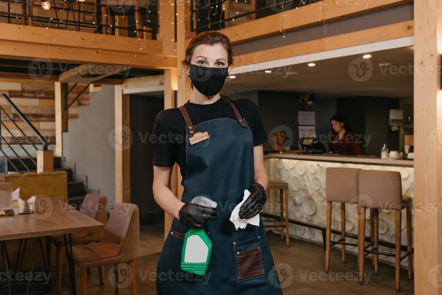 Eine Kellnerin in einer medizinischen Gesichtsmaske hält eine Flasche mit Desinfektionsmittel und einem Lappen in der Hand foto
