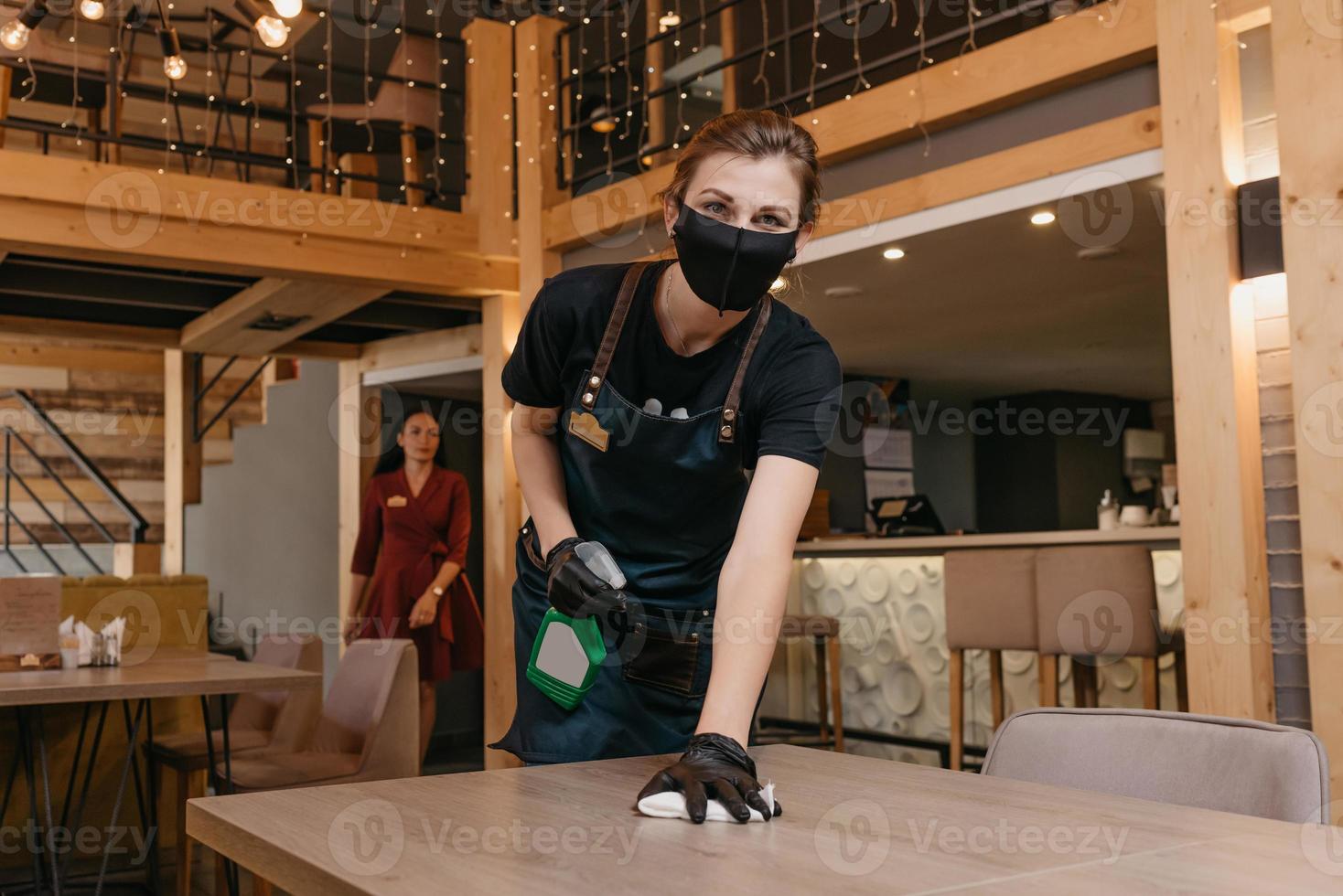 Eine freundliche Kellnerin, die eine schwarze medizinische Gesichtsmaske und medizinische Einweghandschuhe trägt, hält eine Flasche mit Desinfektionsmittel und Reinigungstischen in der Hand foto