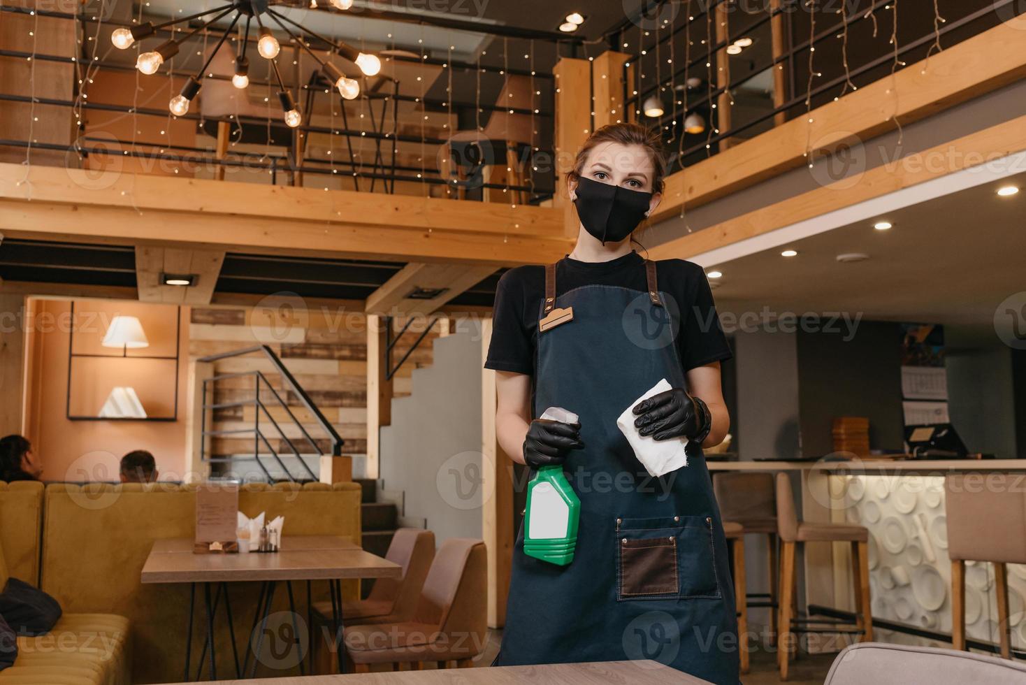 Eine freundliche Kellnerin trägt eine schwarze medizinische Gesichtsmaske und medizinische Einweghandschuhe, die eine Flasche mit Desinfektionsmittel und Reinigungstischen halten foto